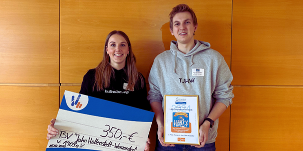 Zwei junge Menschen stehen vor einer Holzwand, eine hält einen großen Scheck über 350 Euro für TSV Jahn Hollenstedt-Wiensdorf, der andere eine Urkunde